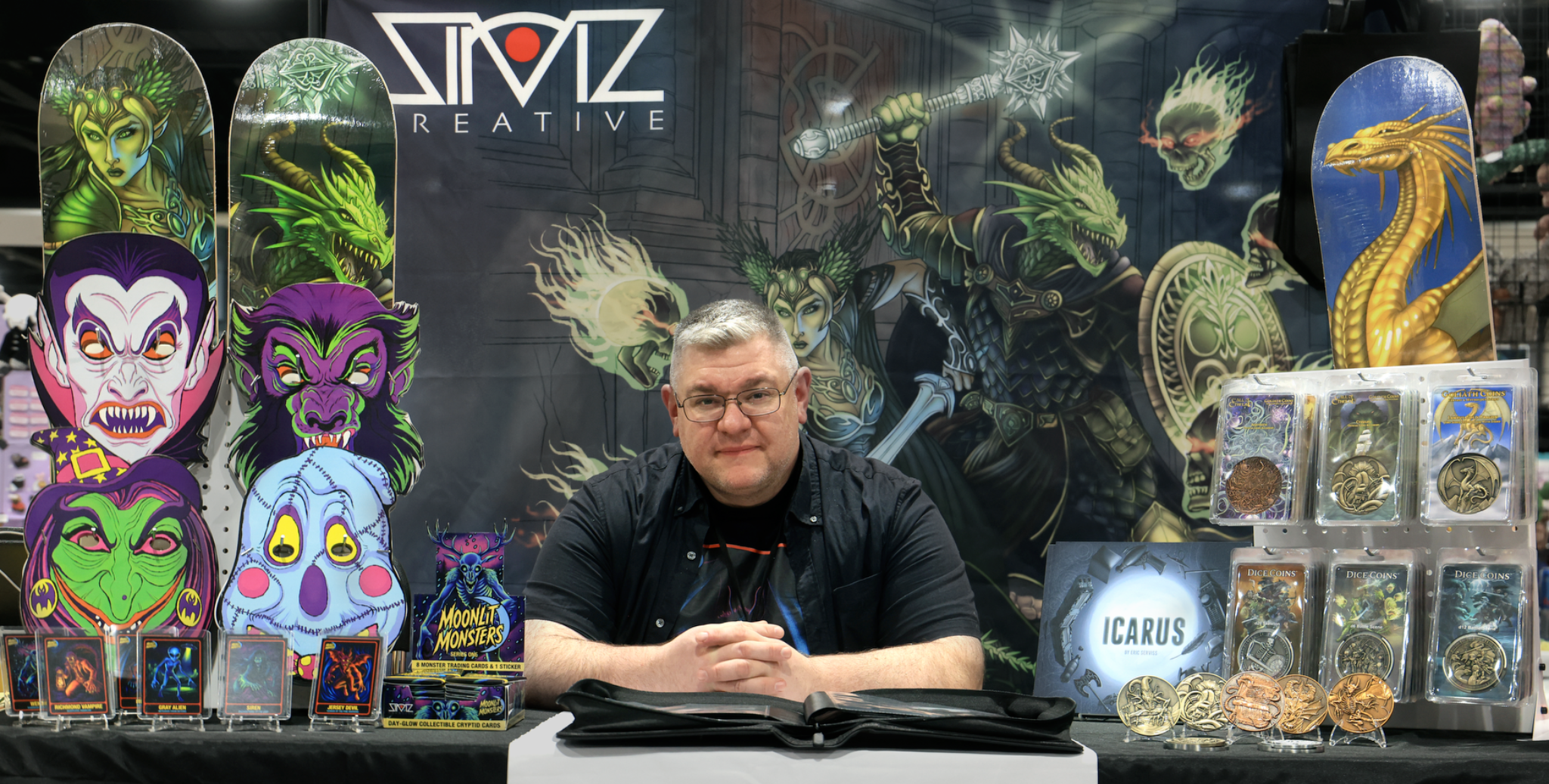 Eric Serviss seated at the SirViz Creative convention display, surrounded by illustrated products, books, cards, and collectible coins